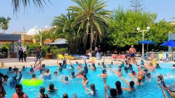 A swimming pool area with people swimming and playing, surrounded by palm trees and umbrellas.