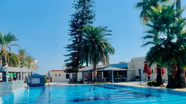 Una piscina con agua azul cristalina, rodeada de palmeras y un edificio de fachada blanca.