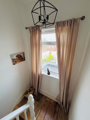Een kamer met een houten trap, een raam met gordijnen, een aan de muur hangend schilderij en een plafondlamp.