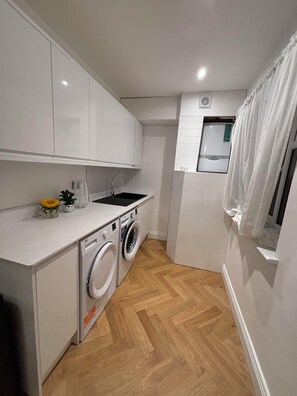 A compact kitchen with white cabinets, a washing machine, and a small window with curtains. - Queens Coast Horizon – Stylish 4-Bedroom Townhouse by Queen's Park | Sleeps 10 (Brighton and Hove)
