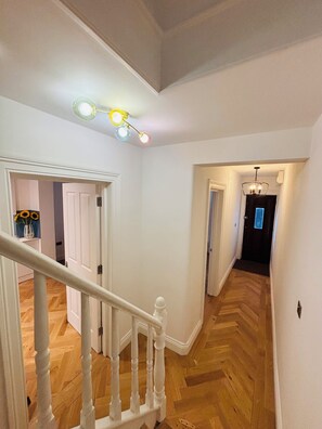 Eine Treppe mit weißen Balustraden, die zu einer Zwischenebene führt, ein Fischgrätparkett und eine Leuchte mit mehreren Glühbirnen.