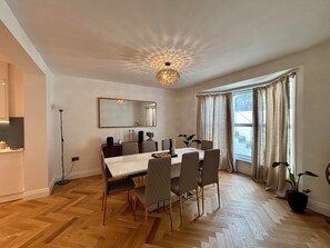 A dining area with a white table, six chairs, a chandelier, a large mirror, and a window with curtains. - Queens Coast Horizon – Stylish 4-Bedroom Townhouse by Queen's Park | Sleeps 10 (Brighton and Hove)