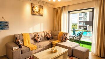 Ein modernes Wohnzimmer mit einer Couch, Hocker und einem Couchtisch. Ein Fenster bietet Blick auf einen Swimmingpool und Gebäude.