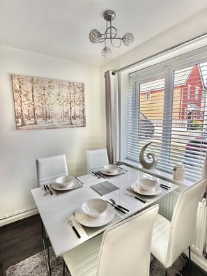 Un coin repas avec une table dressée pour quatre personnes, des chaises blanches, un lustre, une fenêtre avec des stores et un tableau au mur.
