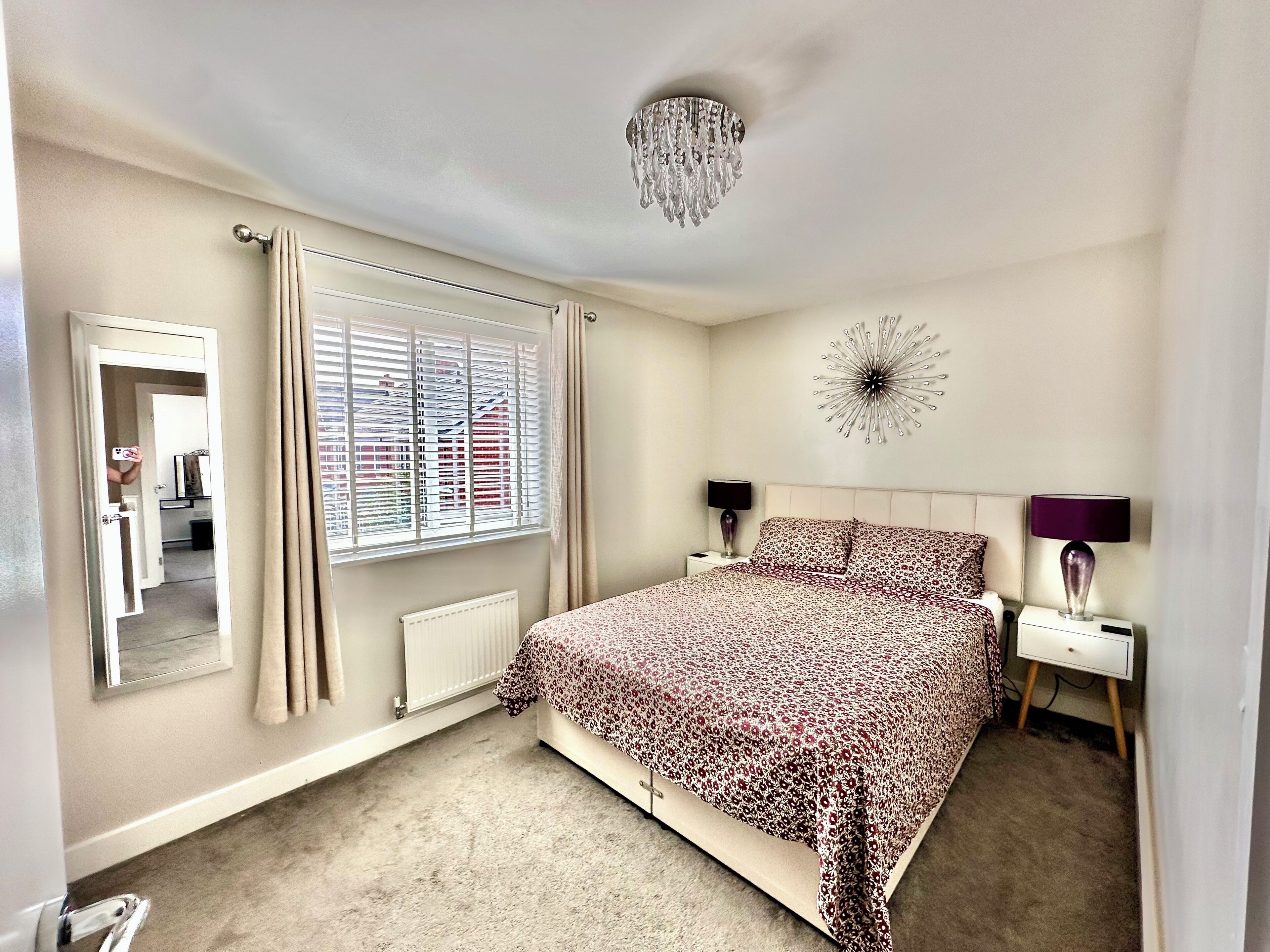 A bedroom with a bed, a nightstand, a mirror, a window with blinds, a radiator, and a wall clock.