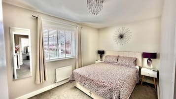 A bedroom with a bed, a nightstand, a mirror, a window with blinds, a radiator, and a wall clock.