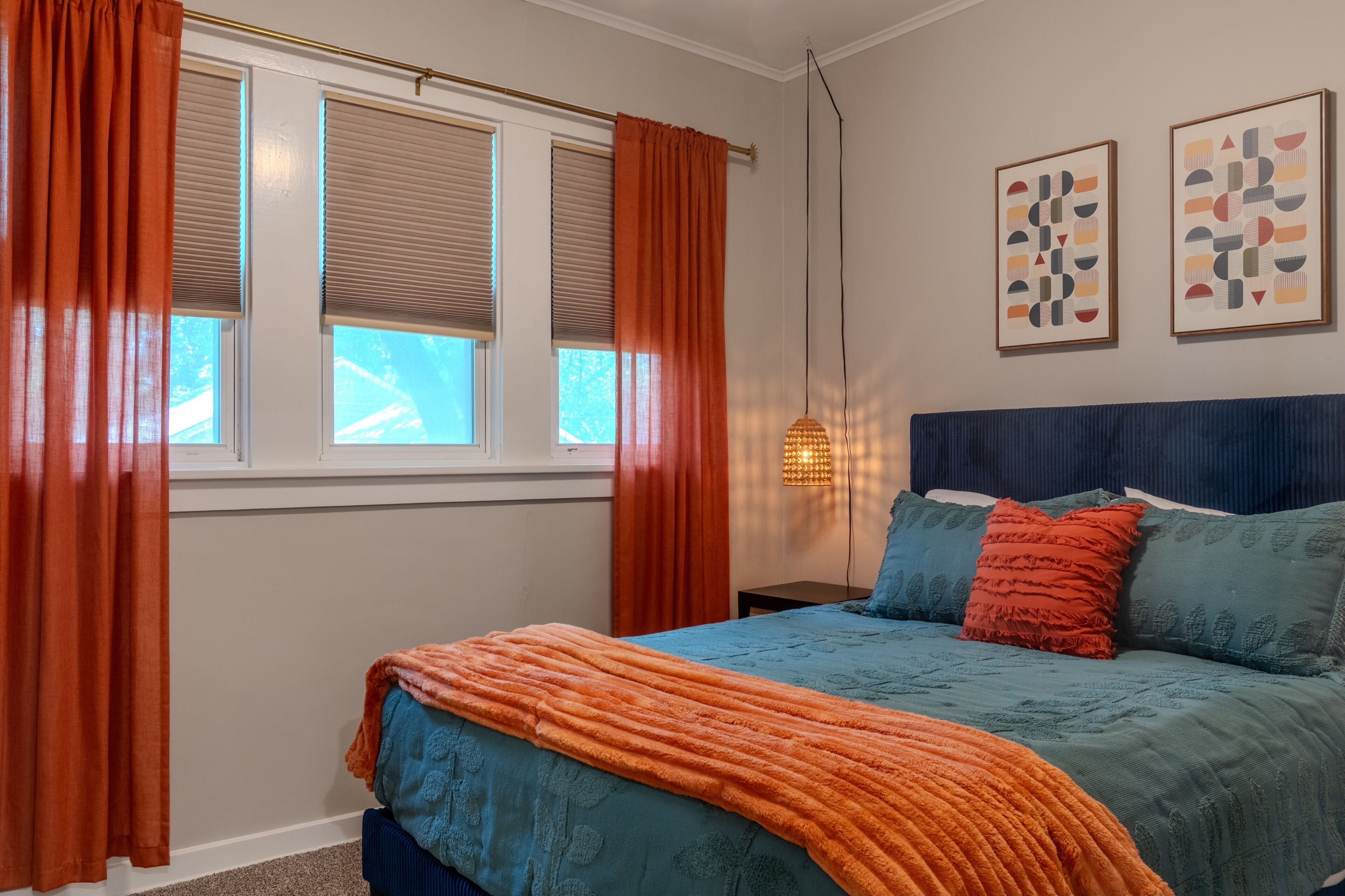 A bedroom with a bed, two windows with blinds, a nightstand, a lamp, and two wall art pieces.