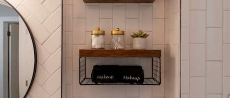 A bathroom with a wooden shelf holding a white towel, a jar with cotton balls, and a potted plant.