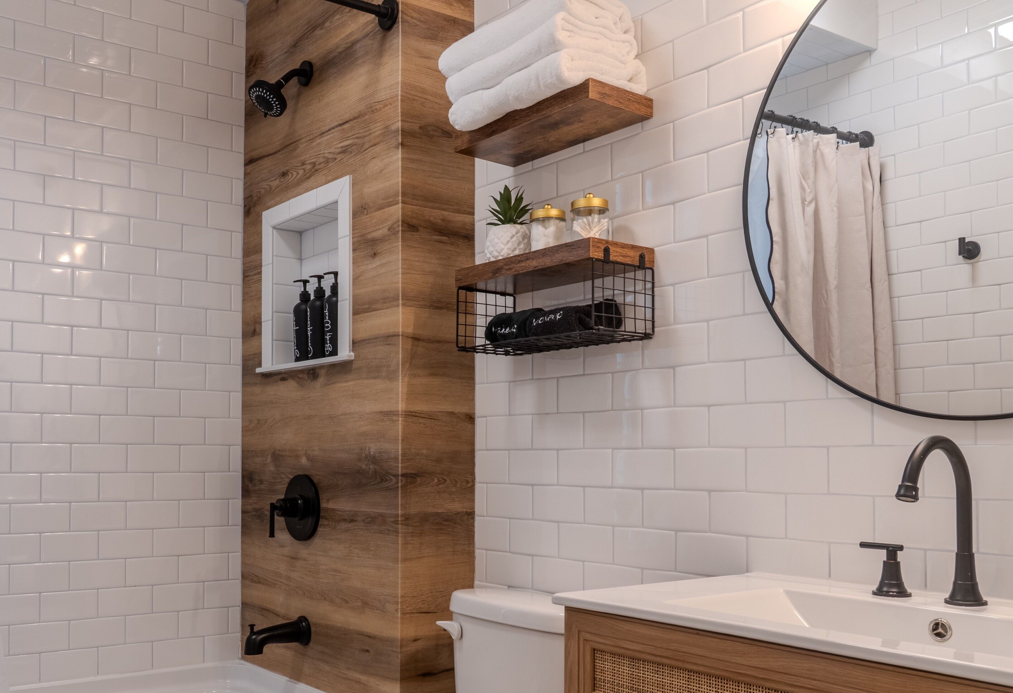 Une salle de bain moderne avec un mur d’accent en bois, un lavabo blanc, des toilettes, une douche et un miroir rond.