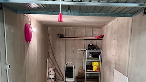 Une pièce avec un sol en béton, un plafond en métal, un luminaire rouge, un balai, une porte blanche et une étagère contenant divers objets.