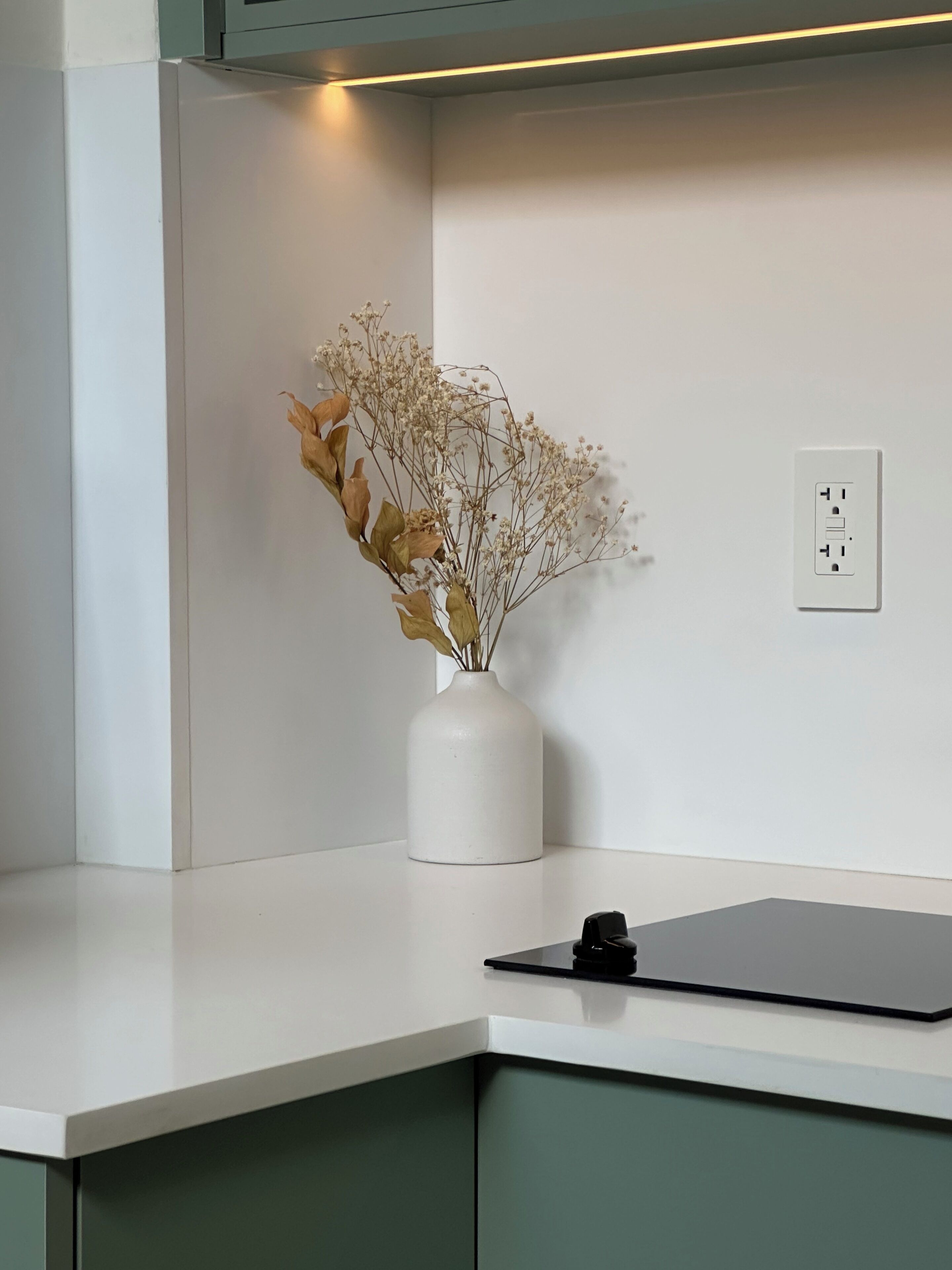 Un vase blanc contenant des fleurs séchées, posé sur un comptoir blanc, un objet noir sur le comptoir et une prise électrique blanche sur le mur.