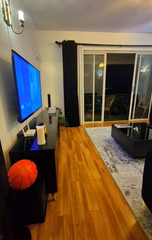 Una sala de estar con una televisión de pantalla plana, un sofá negro, piso de madera, un ventanal que da a un espacio exterior y una pequeña mesa con una planta.