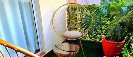 A balcony with two chairs, one with a cushion and one with a woven seat, surrounded by lush green plants and a red pot.