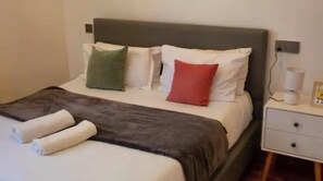 Una cama con cabecera gris, ropa de cama blanca y dos almohadas, una verde y una roja. Hay una mesita de noche blanca con una lámpara y un marco de fotos.