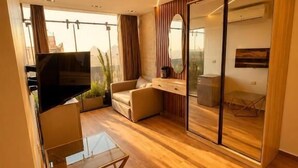 Quarto de hotel moderno com piso de madeira, um sofá, mesa de centro de vidro e uma porta de vidro deslizante para uma varanda.