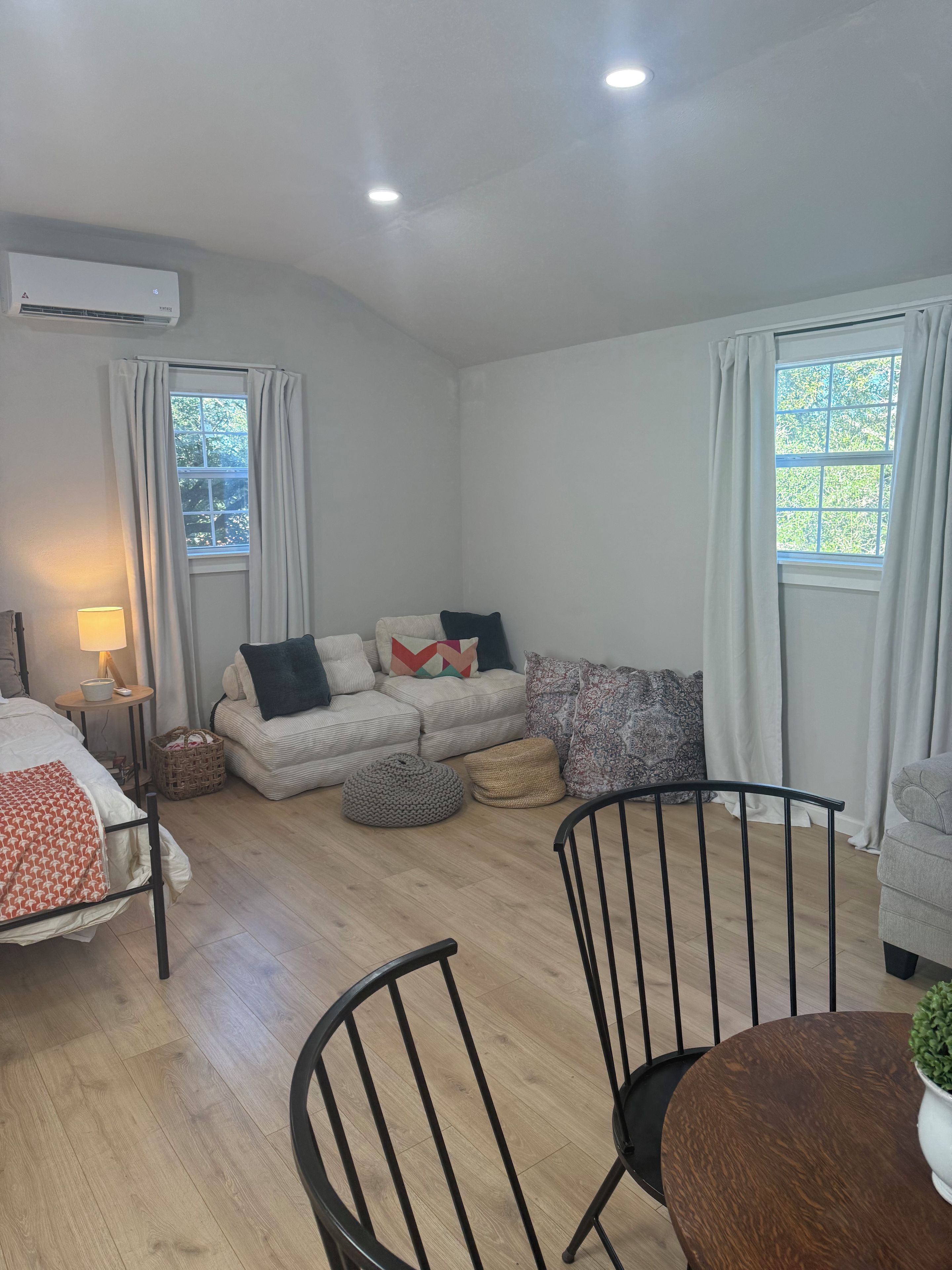 A modern, well-lit room with a bed, a sofa, a dining table, and two windows with curtains.
