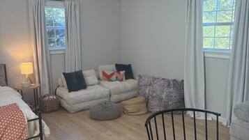 A modern, well-lit room with a bed, a sofa, a dining table, and two windows with curtains.