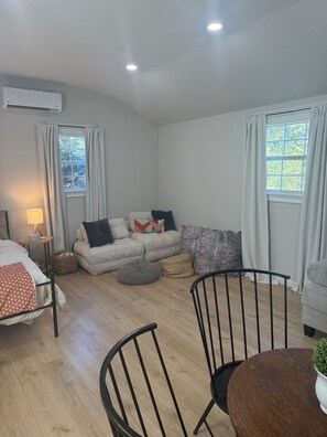 A modern, well-lit room with a bed, a sofa, a dining table, and two windows with curtains. - Lakeside Hideaway, cabin with AC in cozy Brownwood, Walk to Water (Brownwood)