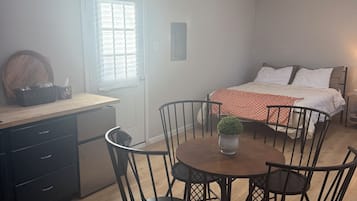 A room with a dining table, chairs, a bed, a window with blinds, and a door.