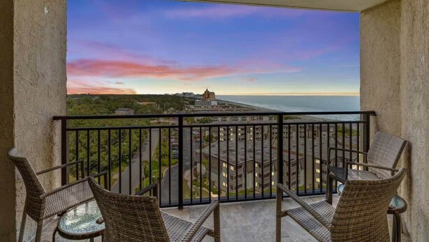 Ein Balkon mit Korbstühlen und einem Tisch, mit Blick auf eine Stadtlandschaft und ein Gewässer bei Sonnenuntergang.