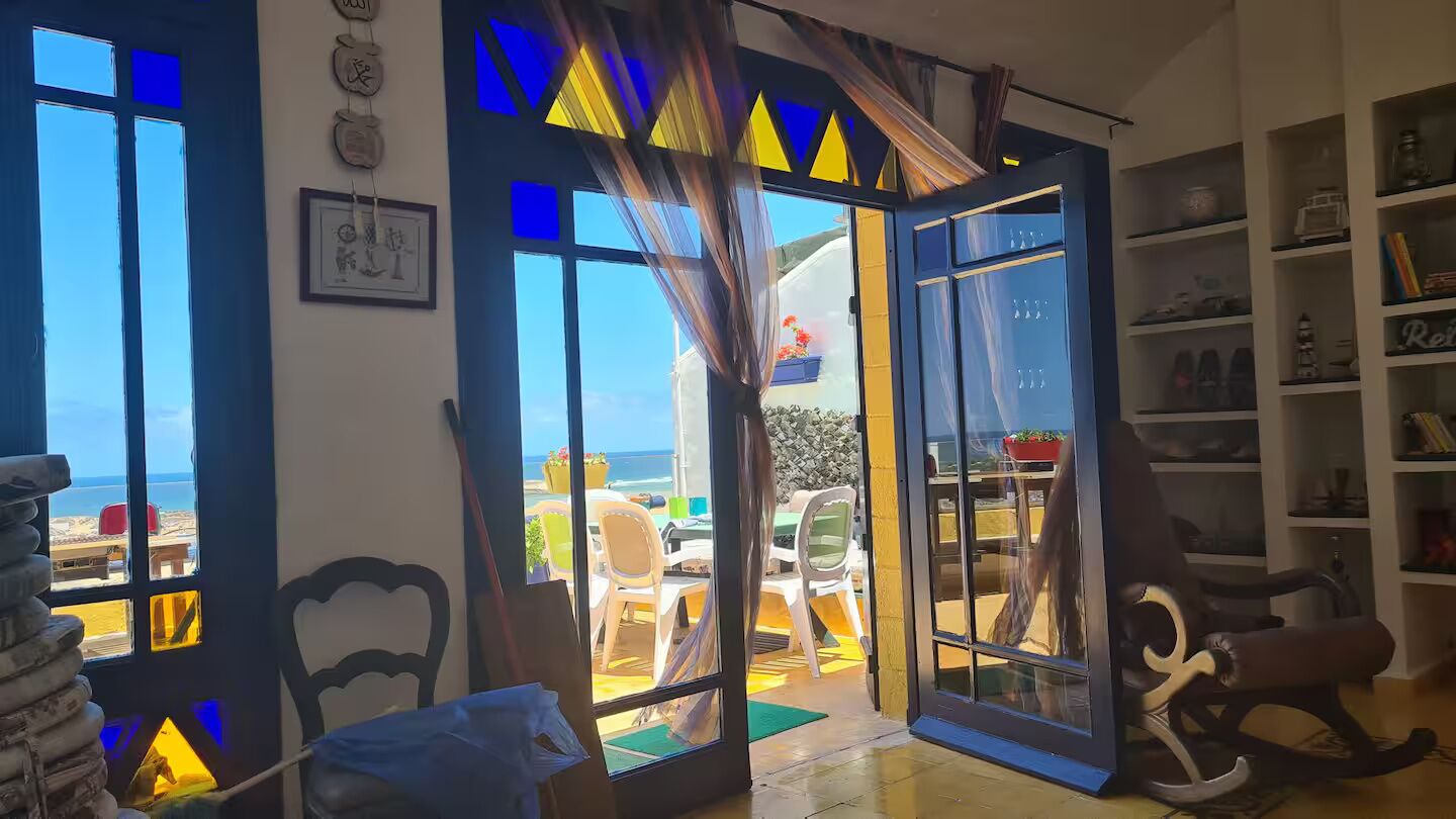 A room with a view of the beach, a dining area with chairs and tables, and a bookshelf.