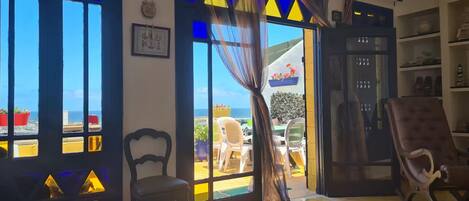 A room with a view of the sea, a chair, a table, and a bookshelf.
