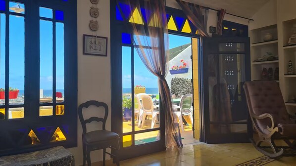 A room with a view of the sea, a chair, a table, and a bookshelf.