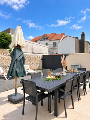 Une terrasse extérieure avec une table dressée pour le repas, entourée de chaises et d’un parasol.