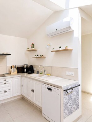 Une cuisine moderne avec des meubles blancs, un lave-vaisselle, une machine à café et un évier. On y trouve également des étagères murales ornées de plantes et un climatiseur fixé au mur.