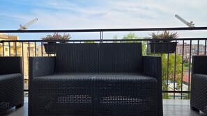 Un balcon avec un canapé et des fauteuils en osier, une balustrade et une vue sur les bâtiments et les arbres.
