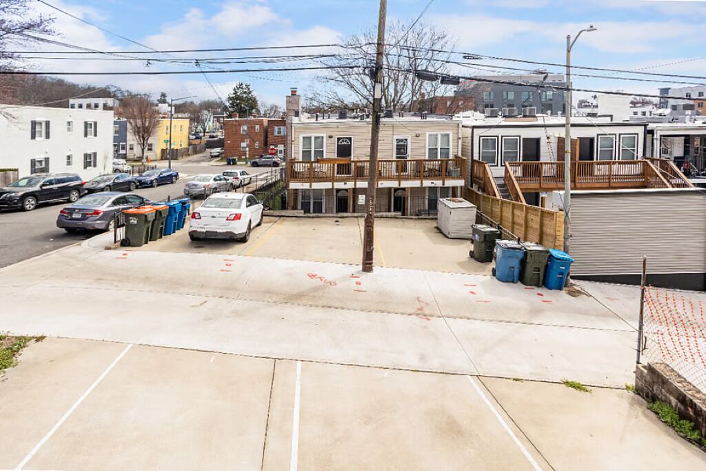 A parking lot with several cars, a building with a wooden deck, and a street with other buildings in the background.