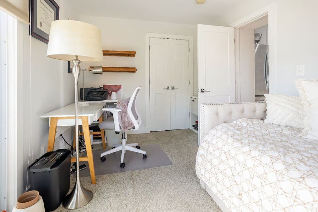A bedroom with a bed, a desk with a chair, a lamp, a door, and a trash bin.