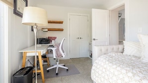 A bedroom with a bed, a desk with a chair, a lamp, a door, and a trash bin. - Lovely 2-bedroom condo with parking near H Street / Union Market (Washington)
