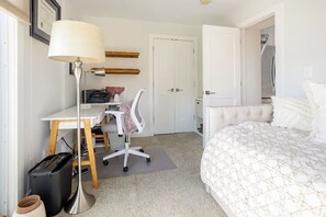 A bedroom with a bed, a desk with a chair, a lamp, a door, and a trash bin. - Lovely 2-bedroom condo with parking near H Street / Union Market (Washington)