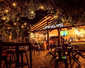 Un área de estar al aire libre con mesas y sillas, una zona de bar con mostrador, y un cartel que dice 'JANGAS'.