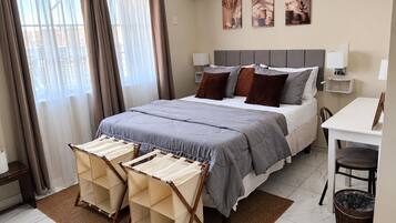 Habitación de hotel con cama, mesitas de noche, escritorio, silla, ventana con cortinas y una obra de arte en la pared.