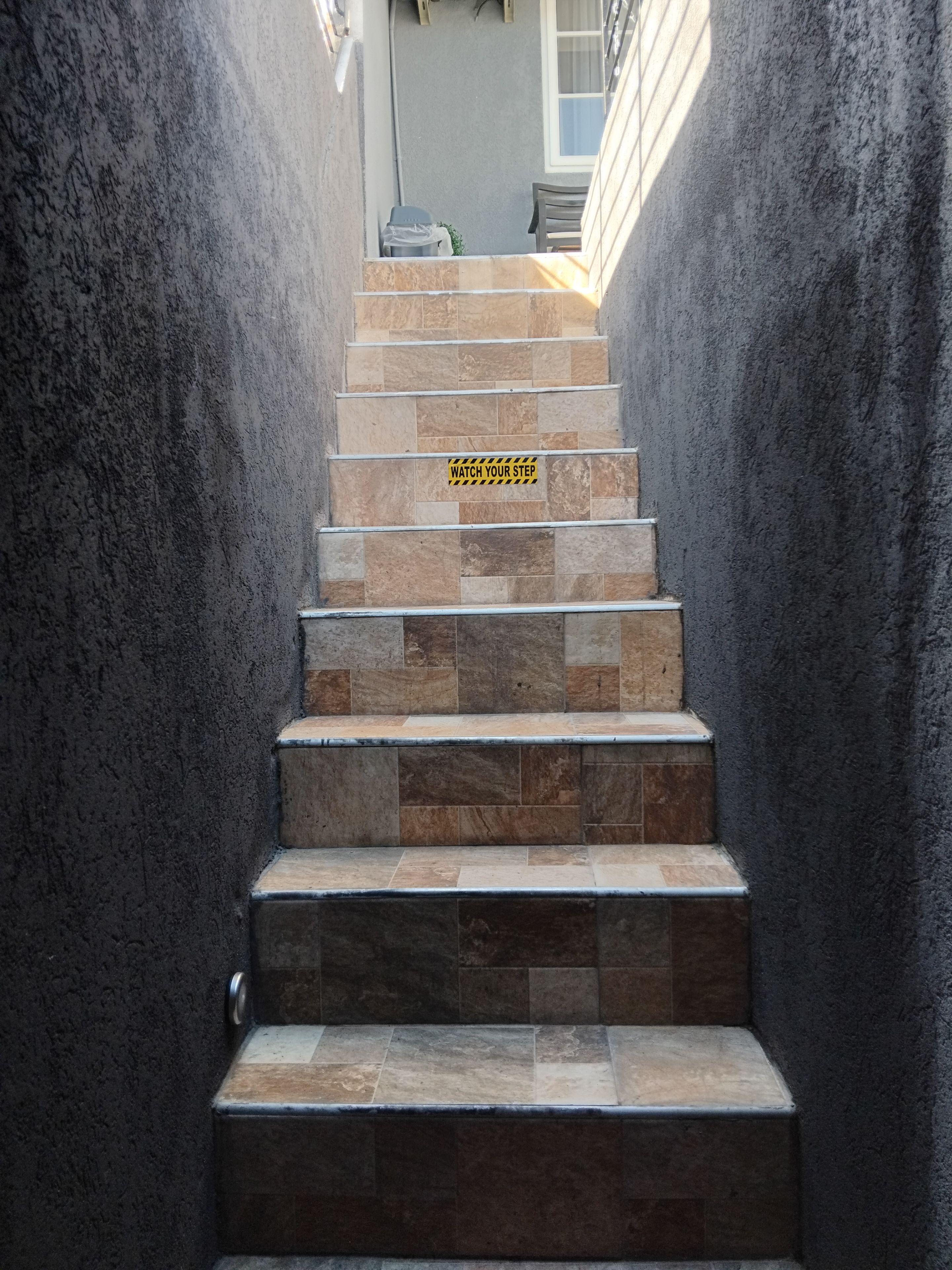 A staircase with a 'WATCH YOUR STEP' sign, leading to a landing with a window and a chair.