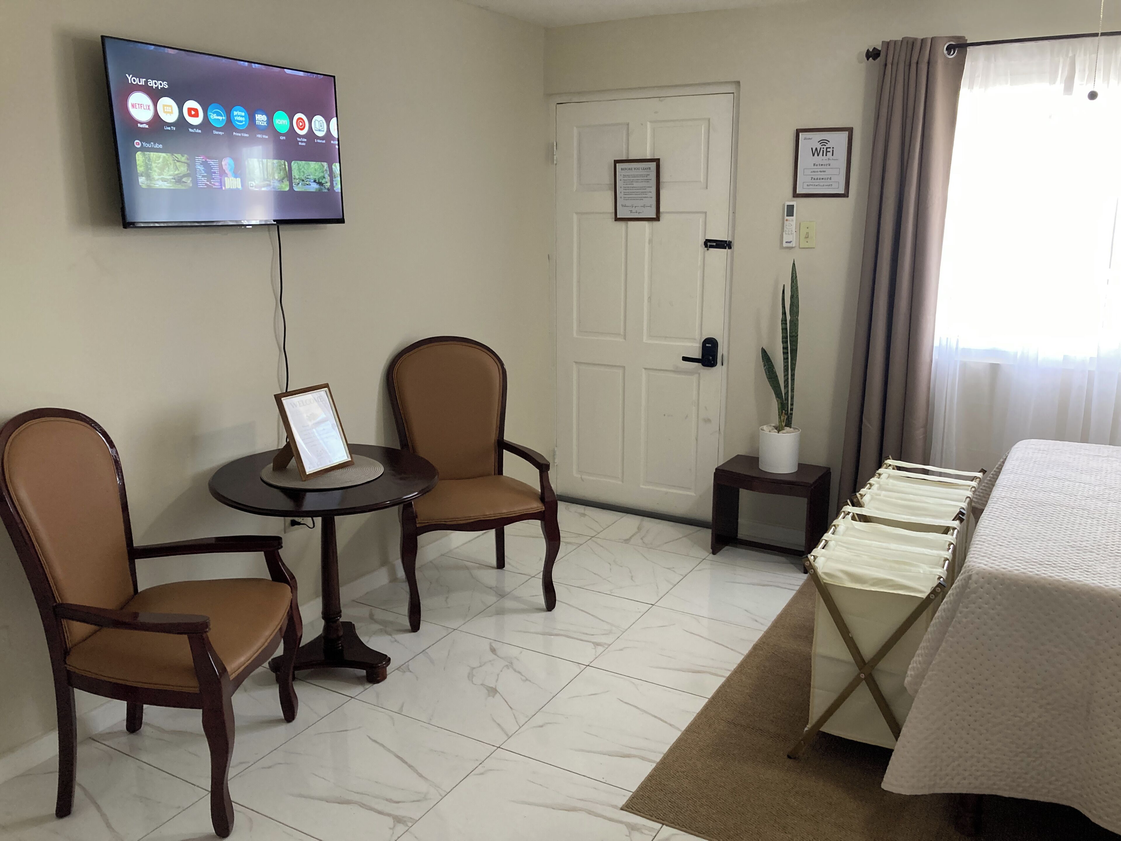 A hotel room with a flat-screen TV mounted on the wall, two wooden chairs, a small table, a bed with a white bedspread, a potted plant, and a door with a sign.