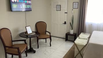 A hotel room with a flat-screen TV mounted on the wall, two wooden chairs, a small table, a bed with a white bedspread, a potted plant, and a door with a sign.