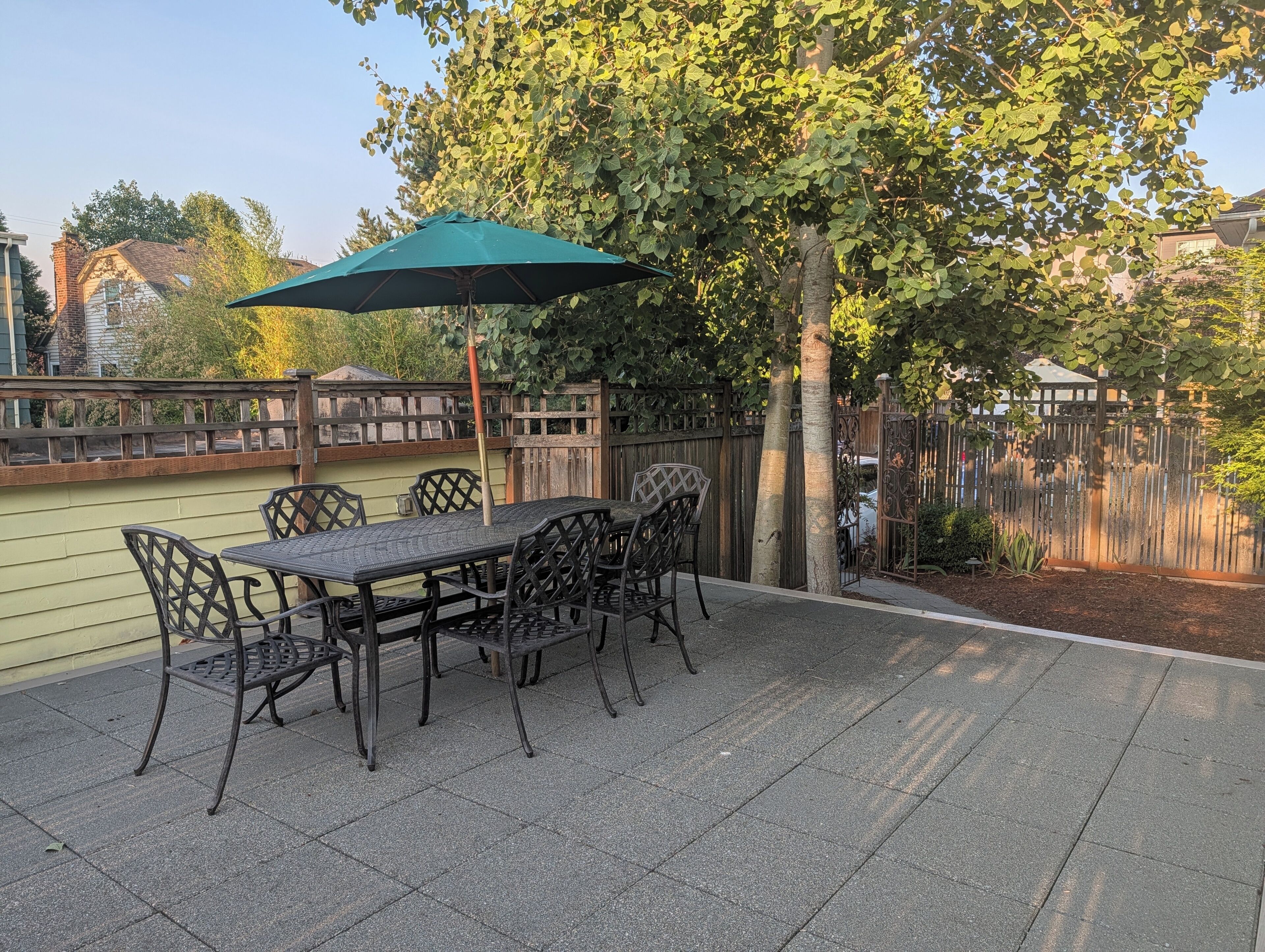 Een terras buiten met een tafel, stoelen, een groene parasol en een houten schutting.