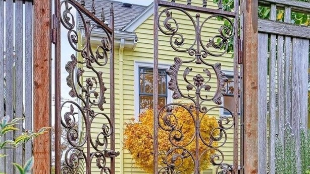 A metal gate with intricate designs, leading to a house with a yellow exterior and a window with a view of a tree with yellow leaves.