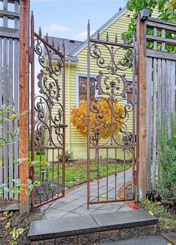 A metal gate with intricate designs, leading to a house with a yellow exterior and a window with a view of a tree with yellow leaves.