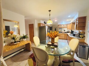A dining area with a glass table, chairs, and a vase with flowers. There is a kitchen in the background with wooden cabinets and a refrigerator.