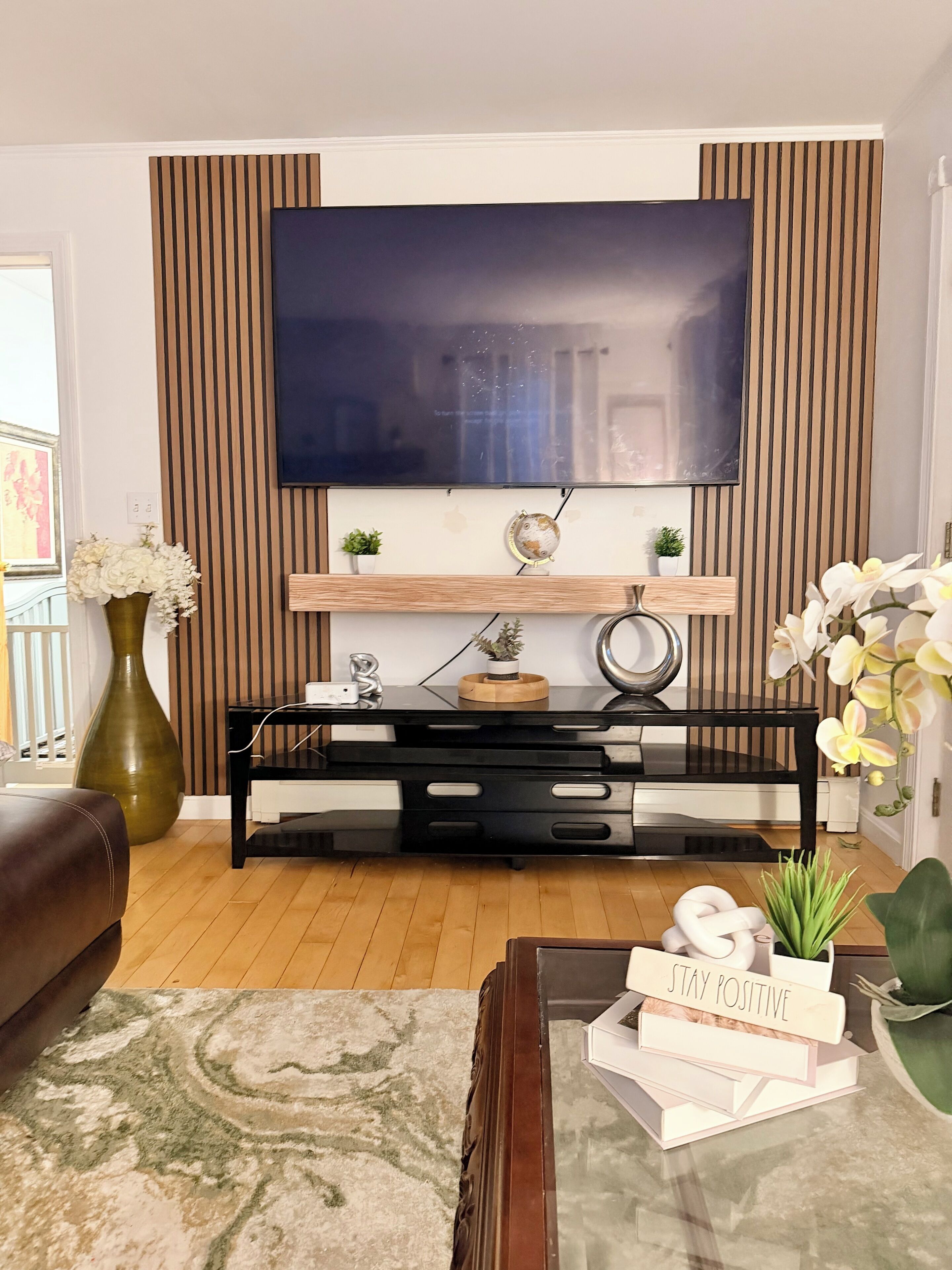A living room with a flat-screen TV mounted on a wall with vertical wooden panels, a black entertainment unit below, a brown leather sofa, a glass coffee table with books and a sign, and a large green vase with white flowers.