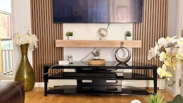 A living room with a flat-screen TV mounted on a wall with vertical wooden panels, a black entertainment unit below, a brown leather sofa, a glass coffee table with books and a sign, and a large green vase with white flowers.