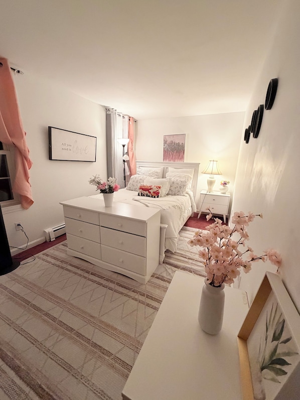 A bedroom with a bed, bedside tables, a dresser, a nightstand, a lamp, a vase with flowers, a framed picture, and a wall-mounted clock.