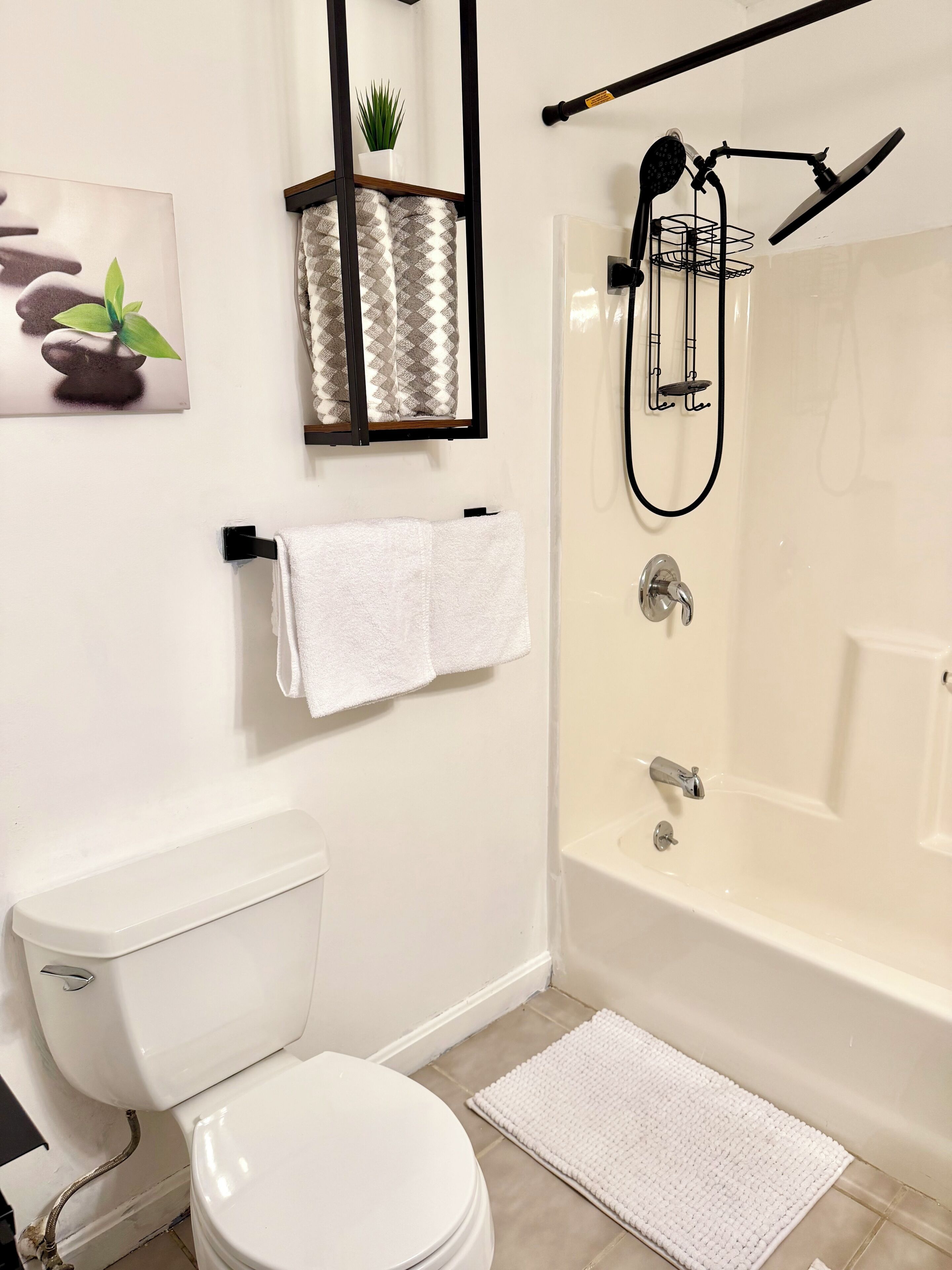 Une salle de bain comprenant des toilettes, une douche avec un pommeau de douche amovible, un porte-serviettes avec une serviette et une petite étagère avec une plante.