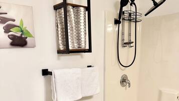 A bathroom with a toilet, a shower with a handheld showerhead, a towel rack with a towel, and a small shelf with a plant.