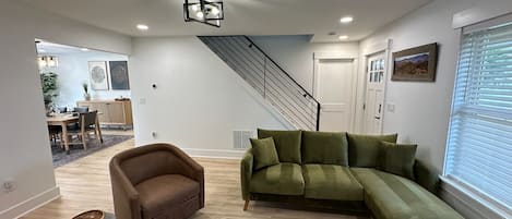 A modern living room with a sectional sofa, a brown armchair, and a patterned rug. A staircase leads to an upper level.