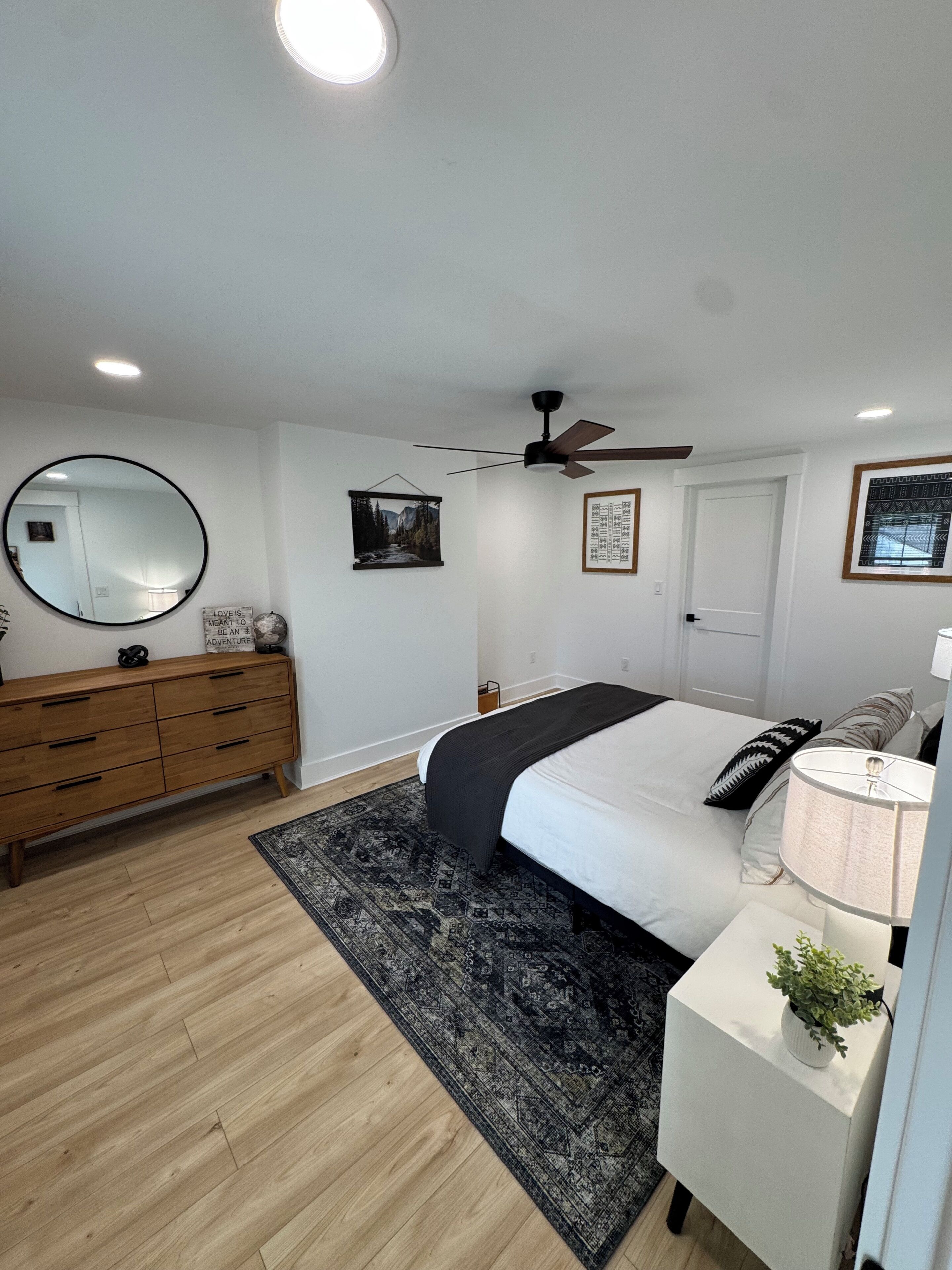 A bedroom with a bed, a bedside table with a lamp, a ceiling fan, a dresser, a rug, and wall decorations.